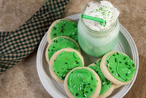 A plate of green frosted cookies accompanied by a mint green milkshake topped with whipped cream and sprinkles, set on a textured surface.