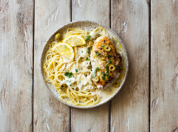 A plate of pasta with grilled chicken topped with cream sauce, olives, and lemon slices on a wooden table