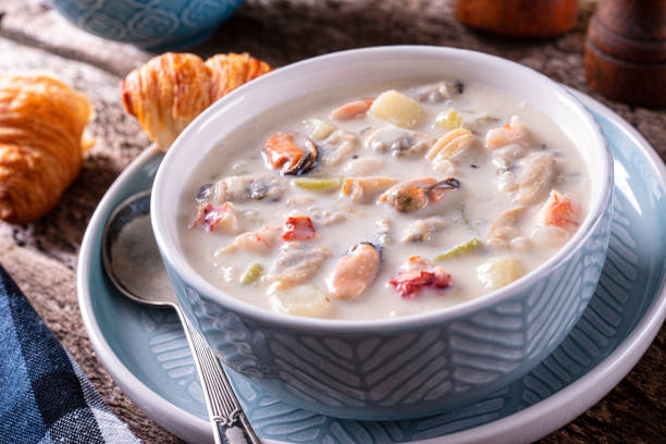 A bowl of creamy seafood chowder with pieces of fish, vegetables, and herbs, accompanied by flaky croissants on a textured surface.