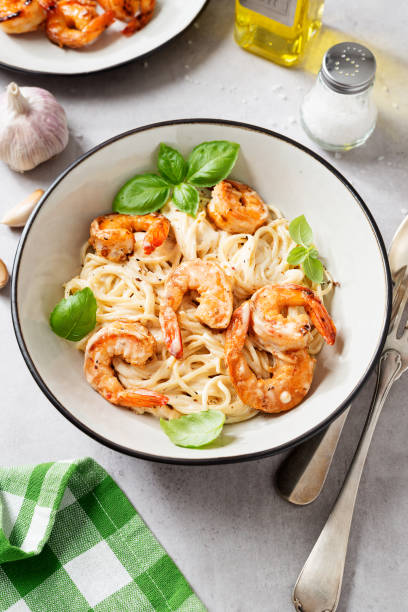 A bowl of shrimp pasta garnished with fresh basil leaves, surrounded by garlic cloves and olive oil.