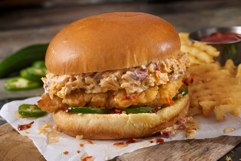 A close-up of a fried chicken sandwich with jalapeños and a golden bun, served with crinkle-cut fries and a small bowl of ketchup.