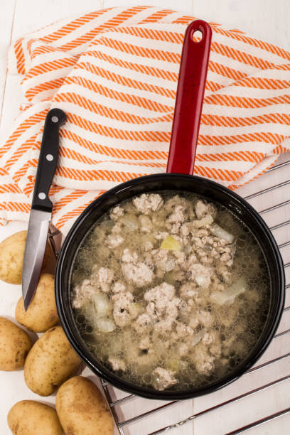 A pot of cooked ground meat in broth, accompanied by fresh potatoes and a knife on a striped kitchen towel.