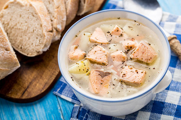 A bowl of salmon soup with chunks of fish and vegetables, beside slices of rustic bread on a blue and white checkered cloth.