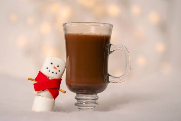 A cozy winter scene featuring a glass of warm chocolate drink next to a cute marshmallow snowman wearing a red scarf.