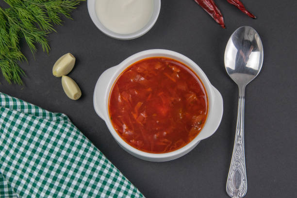 A bowl of red soup with garlic cloves, green herbs, a spoon, and a dish of sour cream on a dark surface.