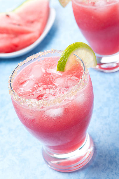 A refreshing watermelon cocktail garnished with a lime slice and served in a glass with a sugared rim, alongside slices of watermelon.