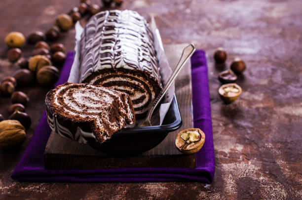 A delicious chocolate and cream striped roll cake served in a black dish with a spoon and surrounded by nuts.