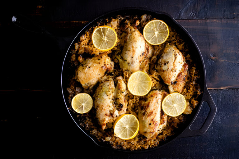 A cast iron skillet filled with golden-brown chicken pieces topped with lemon slices, resting on a bed of rice and spices.