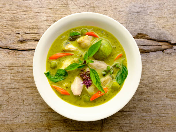 A bowl of green curry with chicken and fresh herbs on a wooden surface.