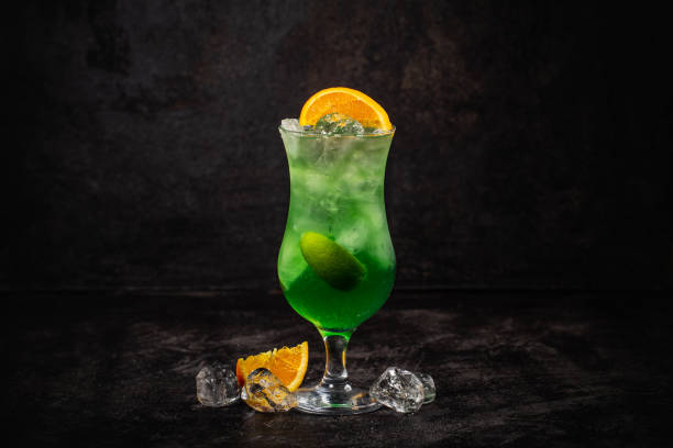 A vibrant green cocktail served in a tall glass, garnished with lime and orange slices, surrounded by ice cubes on a dark background.