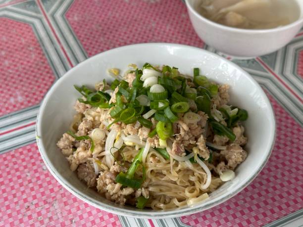 A bowl of noodle dish topped with chopped green onions, served on a patterned tablecloth.