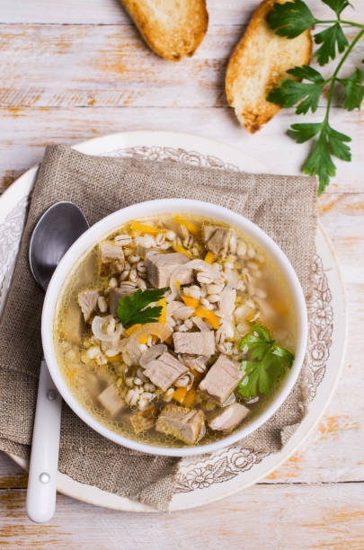 A bowl of hearty soup with pieces of meat and vegetables, garnished with parsley, on a rustic table with slices of toasted bread.