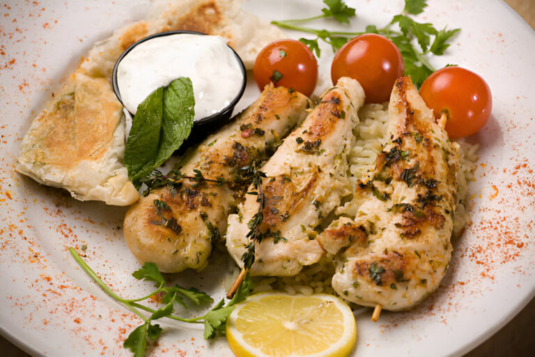 A plate of grilled fish fillets served with rice, cherry tomatoes, a lemon slice, and a side of yogurt sauce with flatbread.