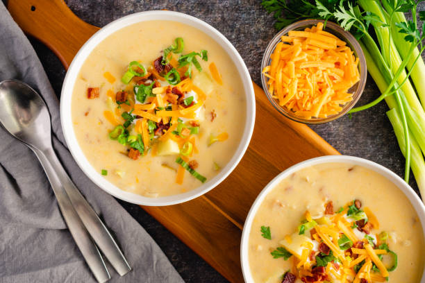 Two bowls of creamy potato soup topped with green onions, cheese, and bacon, alongside a bowl of grated cheese and fresh herbs on a wooden serving board.