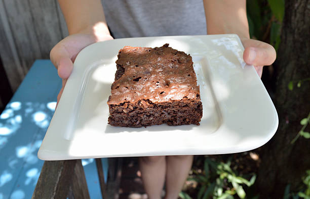 A person holding a slice of brownie on a white plate in an outdoor setting.