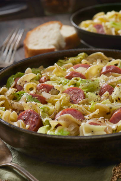 A bowl of pasta with sausage and vegetables, garnished with black pepper, next to slices of bread.