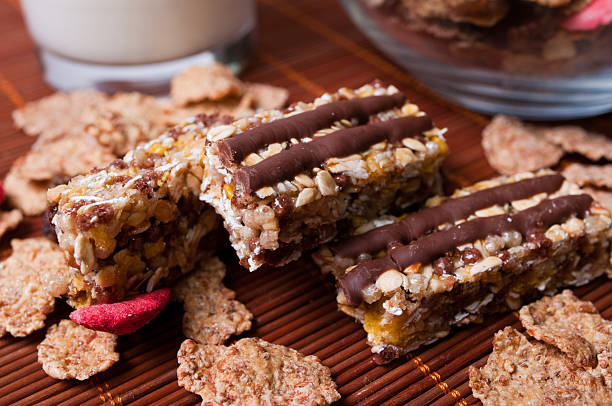 Close-up of chewy granola bars topped with chocolate, surrounded by cereal flakes and a glass of milk in the background.