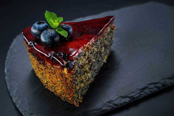 A slice of delicious poppy seed cake topped with berry glaze, blueberries, and a sprig of mint on a black slate plate.