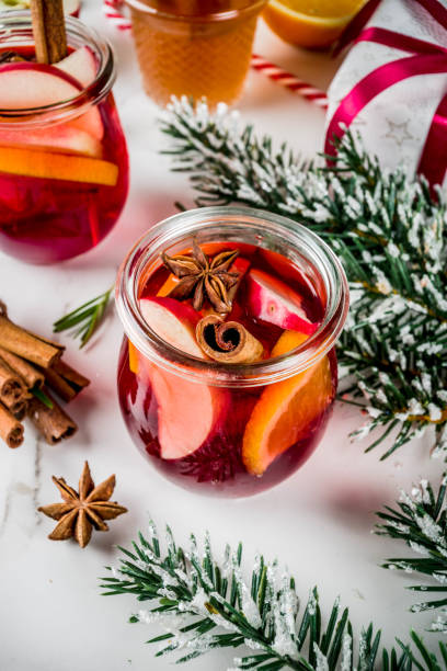 Close-up of a glass jar filled with red beverage, garnished with slices of apple and orange, and decorated with star anise and cinnamon sticks, surrounded by festive greenery.