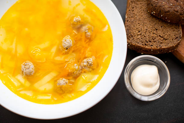 A bowl of soup with meatballs, sliced vegetables, and a side of dark bread and mayonnaise