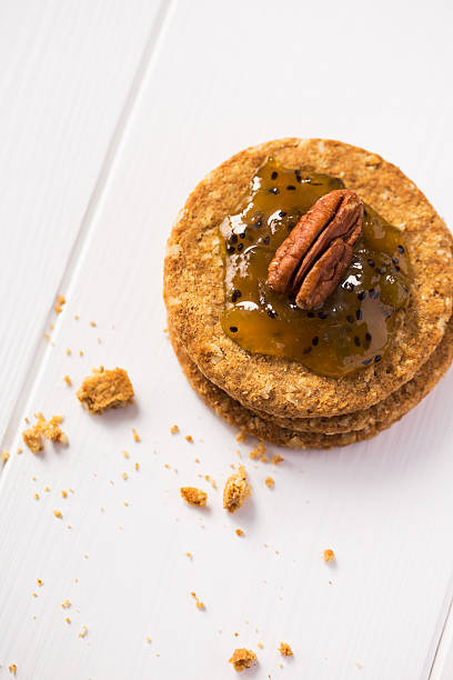 A stack of two round cookies topped with a shiny golden jam and a pecan, placed on a white wooden surface with crumbs around.