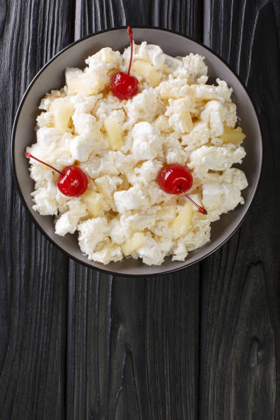 A bowl of cottage cheese with pineapple chunks and cherries on a dark wooden surface.