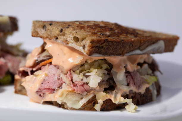 A close-up of a sandwich on a white plate, featuring layers of meat, coleslaw, and pink sauce between slices of toasted brown bread.