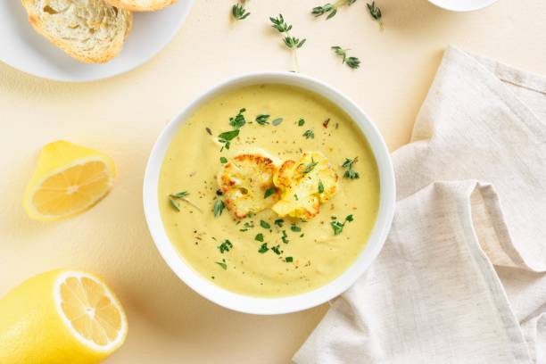 A bowl of creamy soup topped with roasted cauliflower florets, garnished with fresh herbs and accompanied by a slice of bread and lemon on the side.