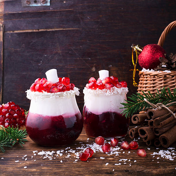 Two glass desserts with pomegranate seeds and coconut on a wooden table, surrounded by pine branches, cinnamon sticks, and a festive basket.