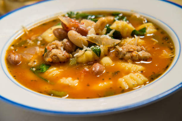 A bowl of hearty vegetable and meat soup with gnocchi and beans, garnished with cheese and herbs.