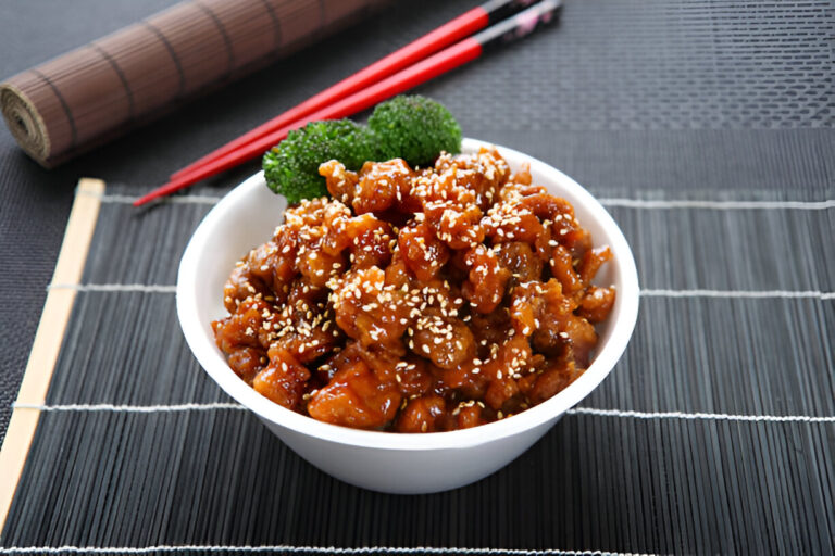 A bowl of sesame chicken garnished with sesame seeds and broccoli on a textured black surface.