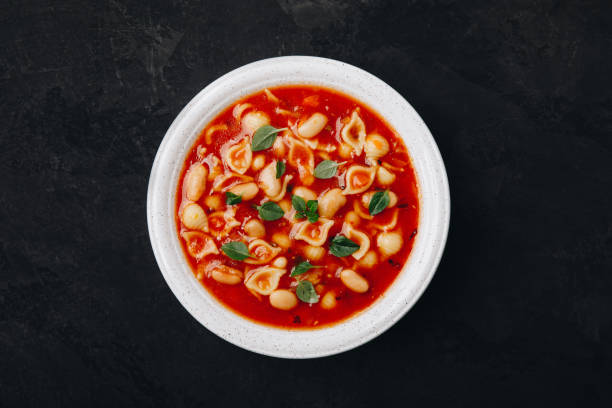 A bowl of pasta soup with red broth and garnished with fresh herbs.