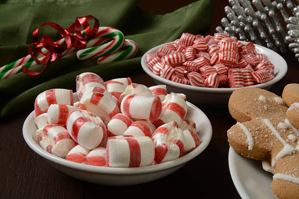 Bowl of red and white peppermint candies, bowls of hard candies, and gingerbread cookies on a wooden table with a green cloth.