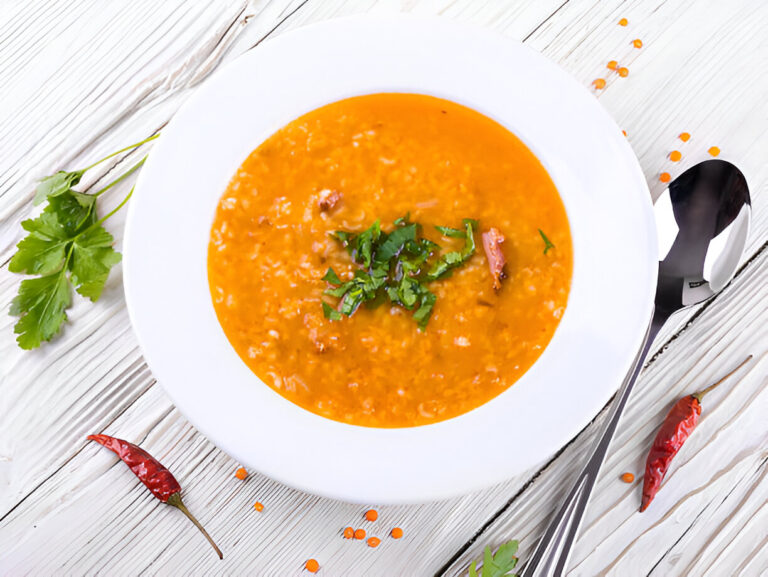 A bowl of rich orange soup garnished with fresh cilantro, placed on a wooden surface with scattered red lentils and dried chilies.