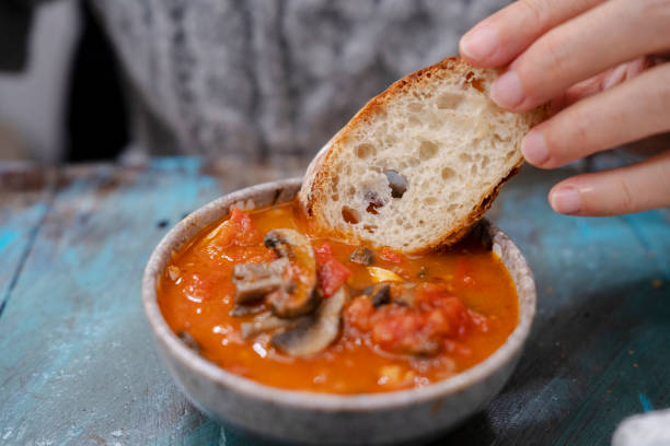 A hand dipping a slice of bread into a bowl of tomato and mushroom soup on a rustic wooden table.