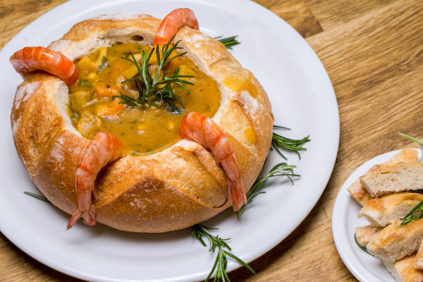 A bowl of soup served in a bread bowl, garnished with shrimp and fresh herbs on a wooden table.