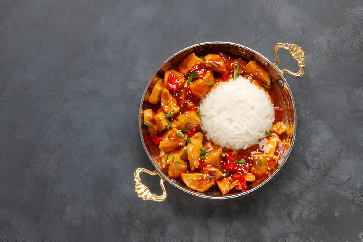 A steaming bowl of chicken in a sweet and spicy sauce served with a mound of fluffy white rice, garnished with green herbs in a decorative metal bowl.