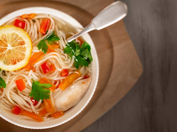 A bowl of chicken noodle soup with vegetables, garnished with a lemon slice and fresh parsley, placed on a wooden surface.