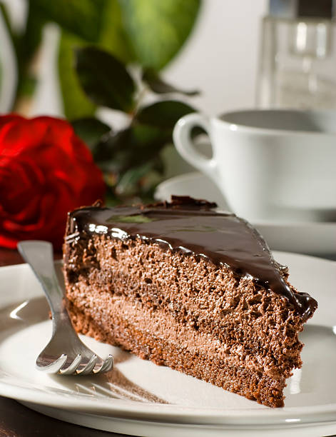 A slice of decadent chocolate cake on a white plate with a fork, accompanied by a red rose and a cup of coffee in the background.