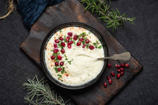 A bowl of creamy white dip garnished with pomegranate seeds, herbs, and spices, served on a wooden board.