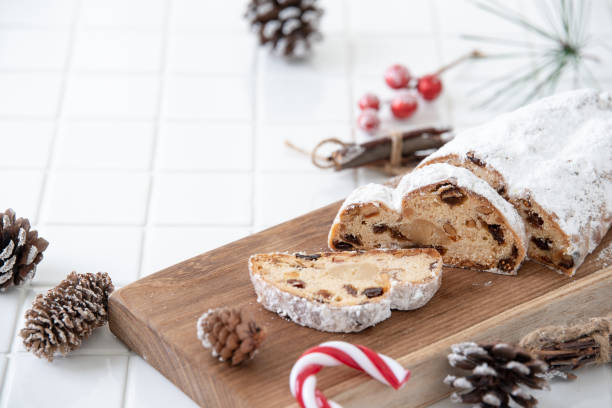 A sliced stollen cake dusted with powdered sugar on a wooden platter, surrounded by pine cones and red berries.