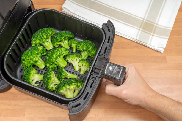 Fresh broccoli florets in an air fryer basket with a hand reaching to pull it out.