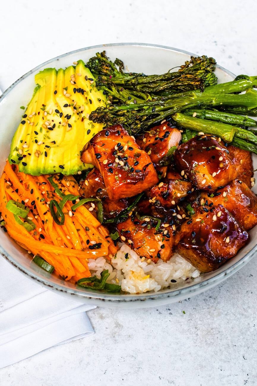 A vibrant bowl of rice topped with glazed salmon, sliced avocado, carrot strips, and green asparagus, garnished with sesame seeds and green onions.