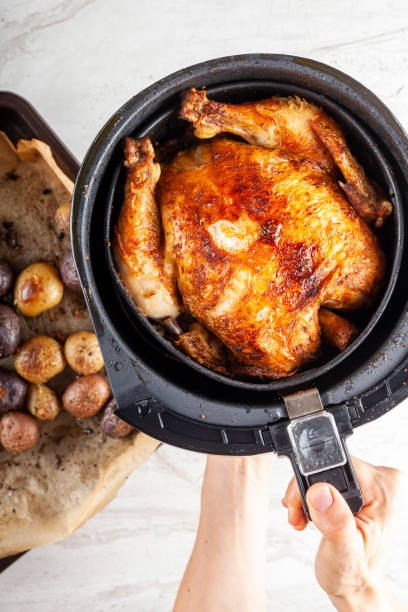 A perfectly cooked whole roasted chicken being lifted from an air fryer by a hand, with roasted potatoes beside it.