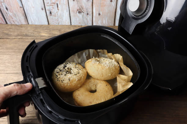 A hand opening an air fryer containing three freshly cooked bagels on parchment paper.