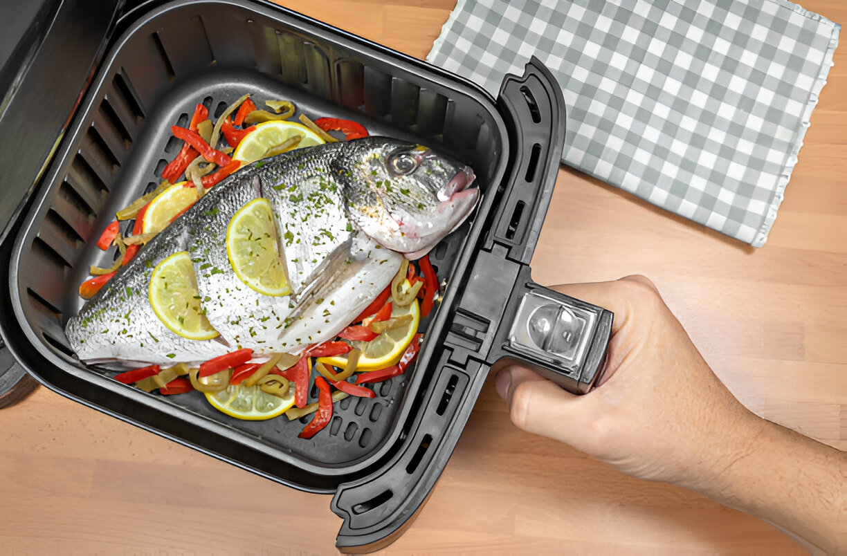 A hand holding an air fryer basket containing a whole fish with lemon slices and colorful bell peppers.