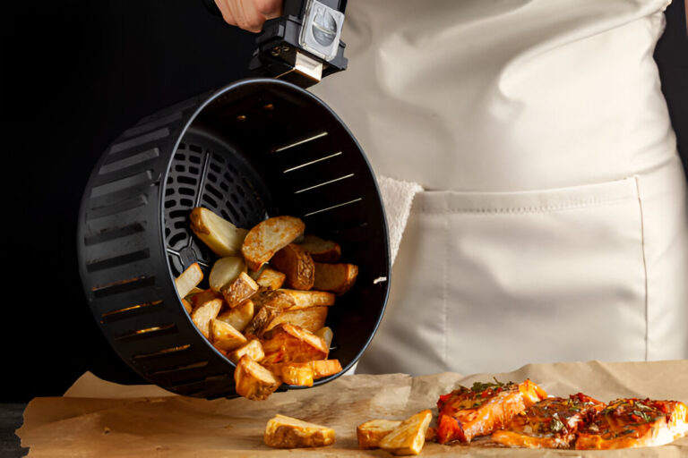 A person pouring golden-brown cooked potato wedges from an air fryer basket onto a wooden surface with a piece of chicken beside it.