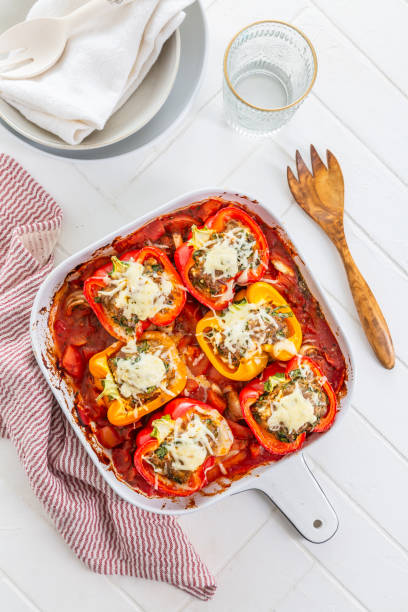 A baking dish filled with stuffed bell peppers topped with melted cheese, surrounded by utensils and a plate.