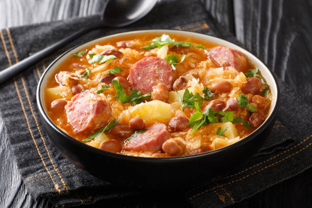 A bowl of hearty stew with sausages, beans, and herbs on a black wooden table.