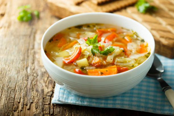 A bowl of vegetable soup with carrots, peppers, and herbs on a wooden table.